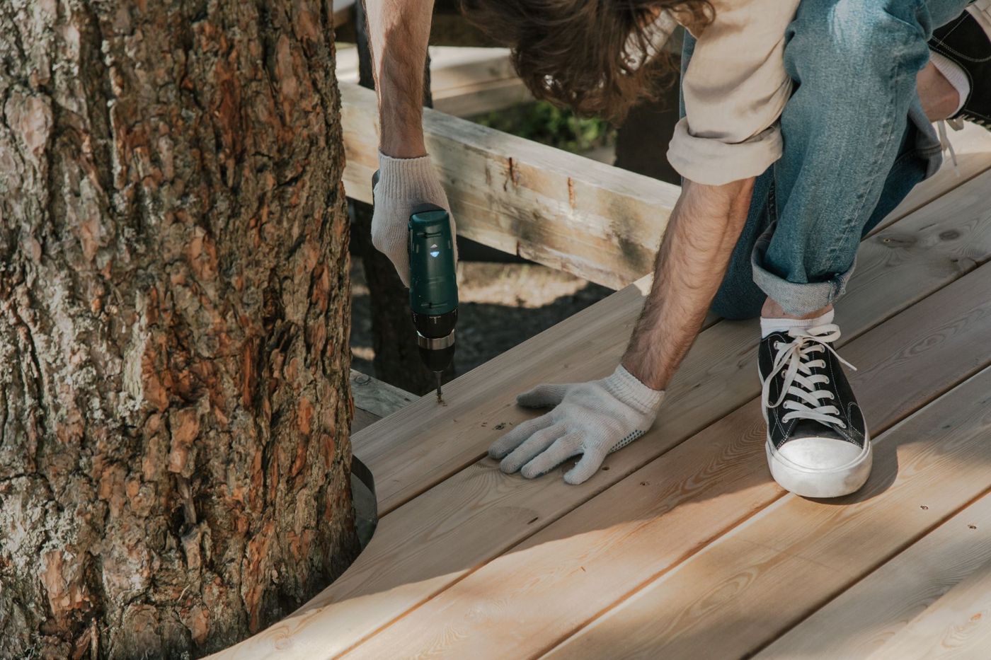 Gloved hands driving a screw into light wood deck boards next to a tree trunk