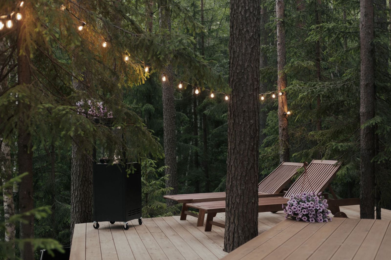Light wood deck built around tall evergreen trunks, with two wooden sun loungers and warm string lights overhead at dusk