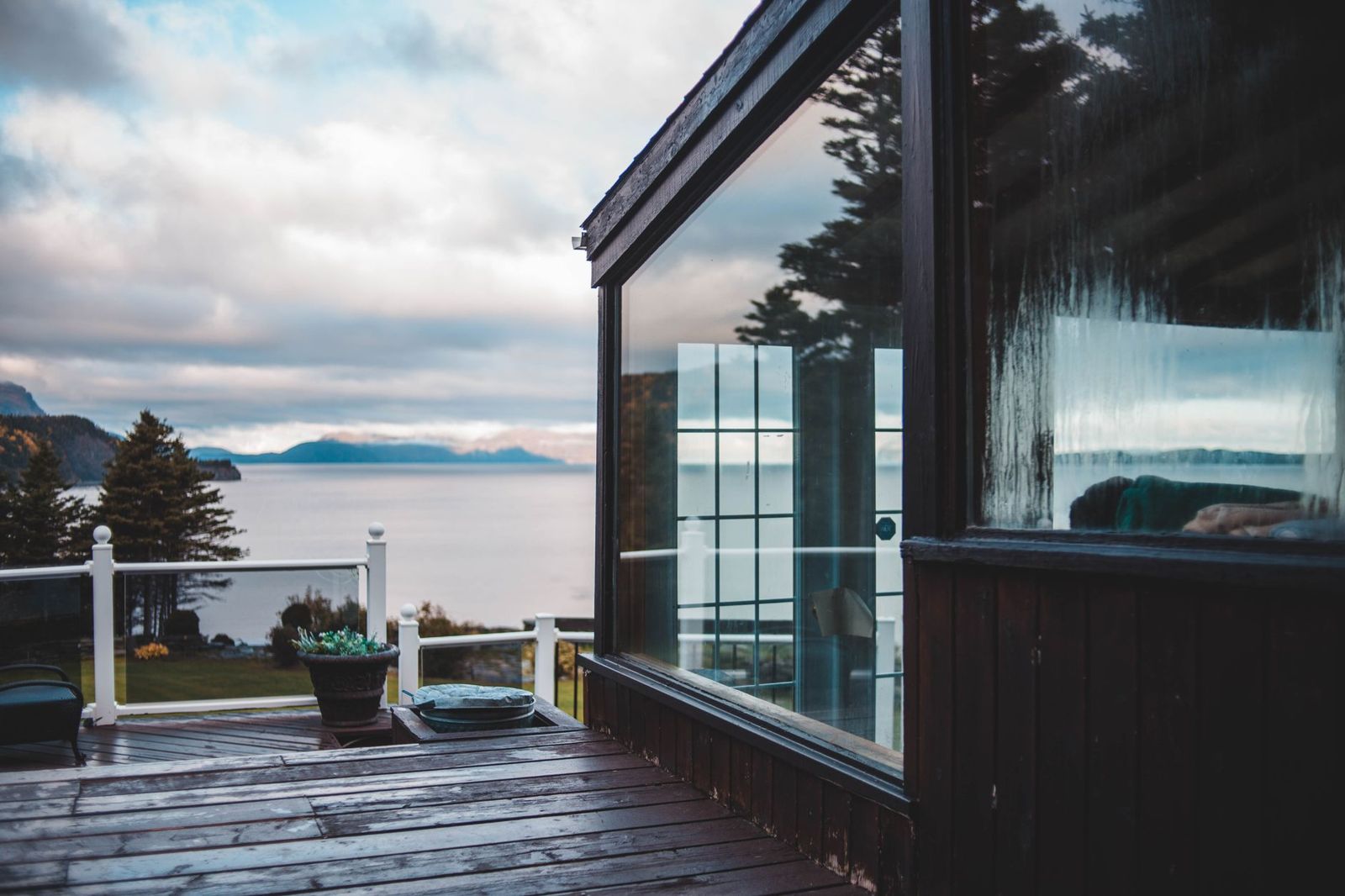Dark wood deck beside a glass-walled sunroom, with white posts and glass railing panels looking out over a wide bay and hills