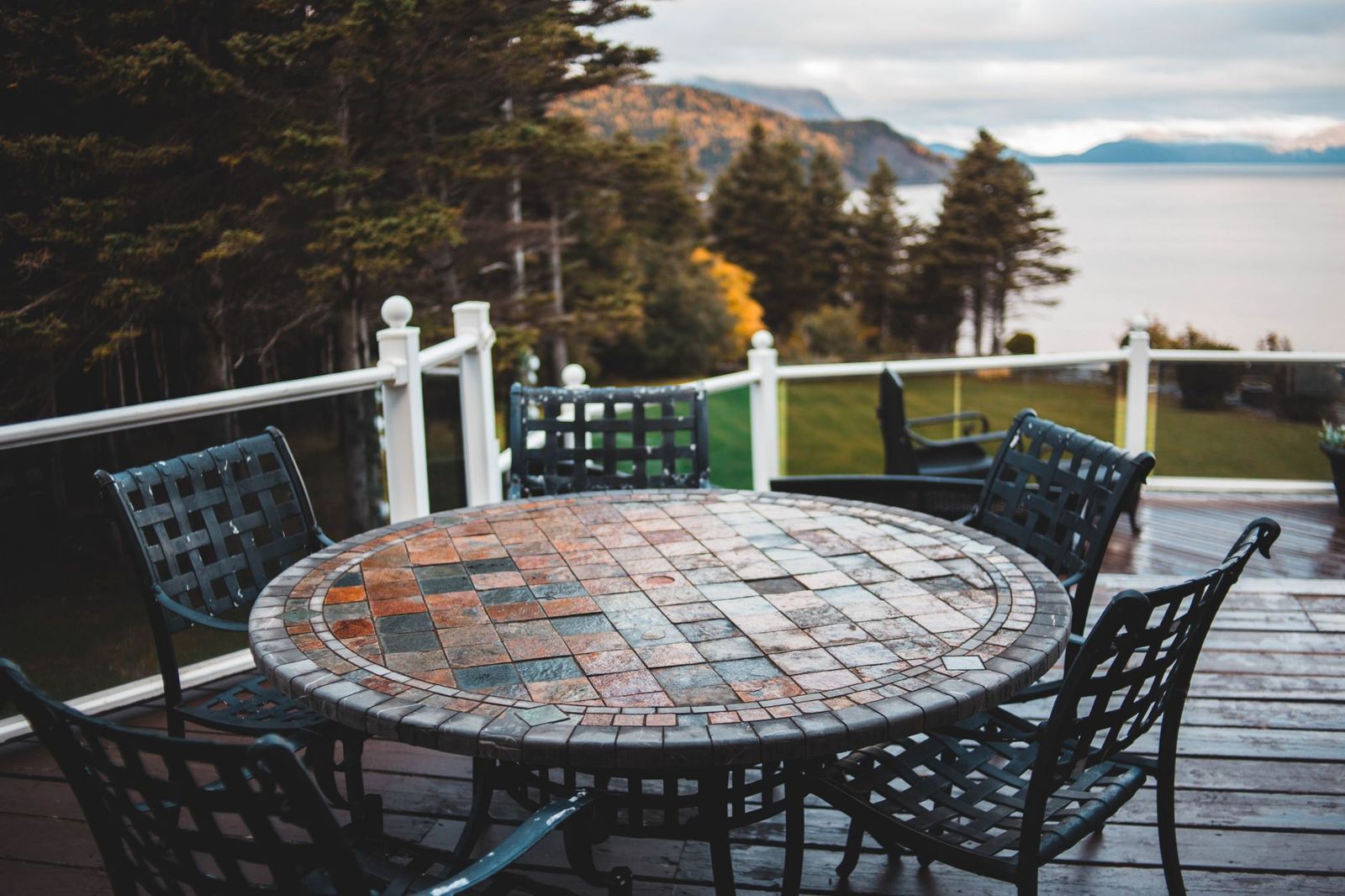 Rain-wet dark wood deck with white railing posts, a round mosaic-top table and black metal chairs above a forested bay