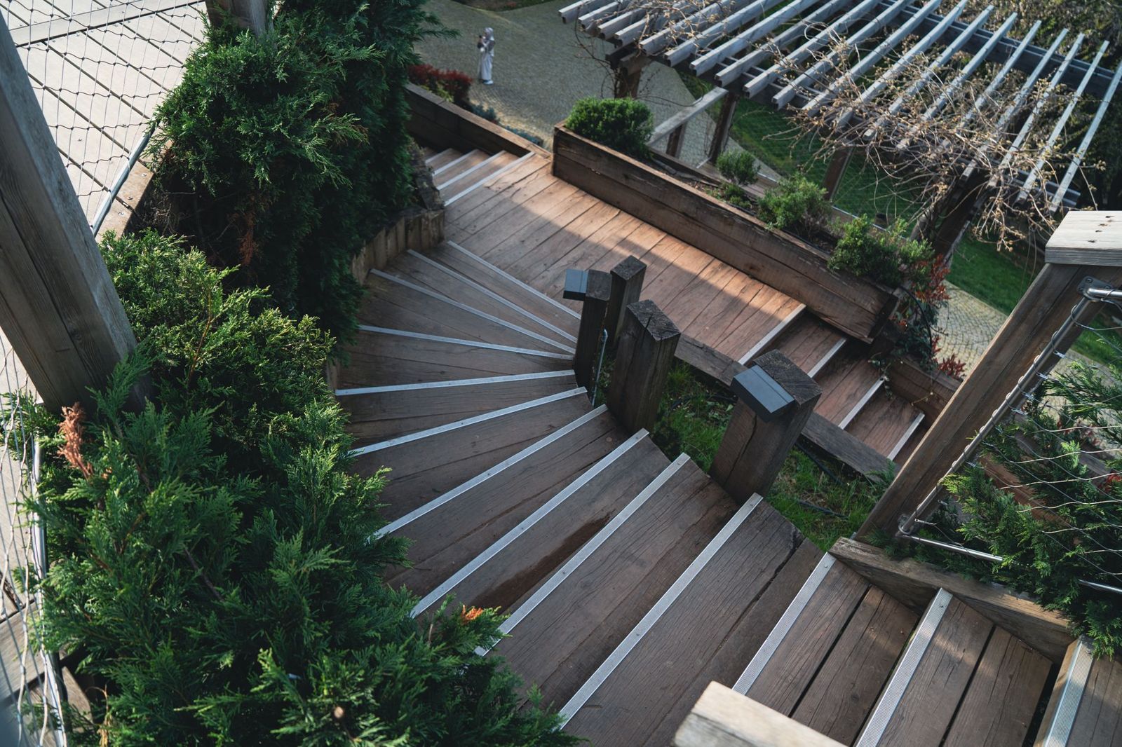 Winding outdoor wooden staircase with light-colored stair nosing, square posts and planters, descending through evergreen shrubs