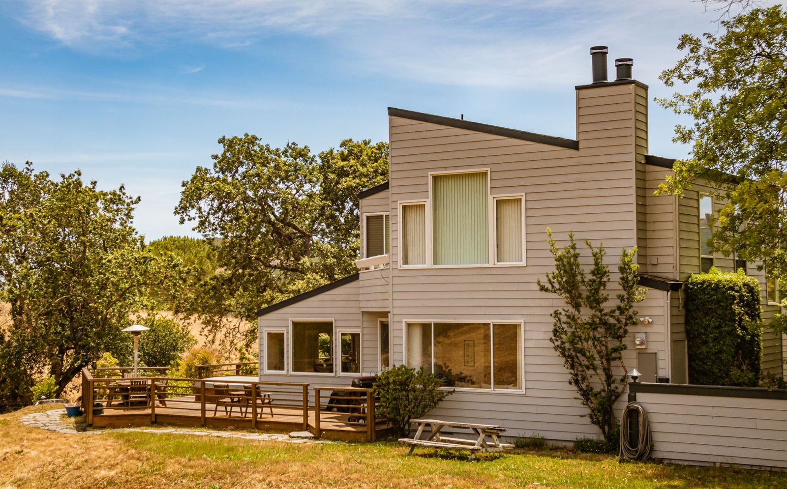 Two-storey house with grey lap siding and a ground-level wood deck with railing and patio table on a sunny lawn